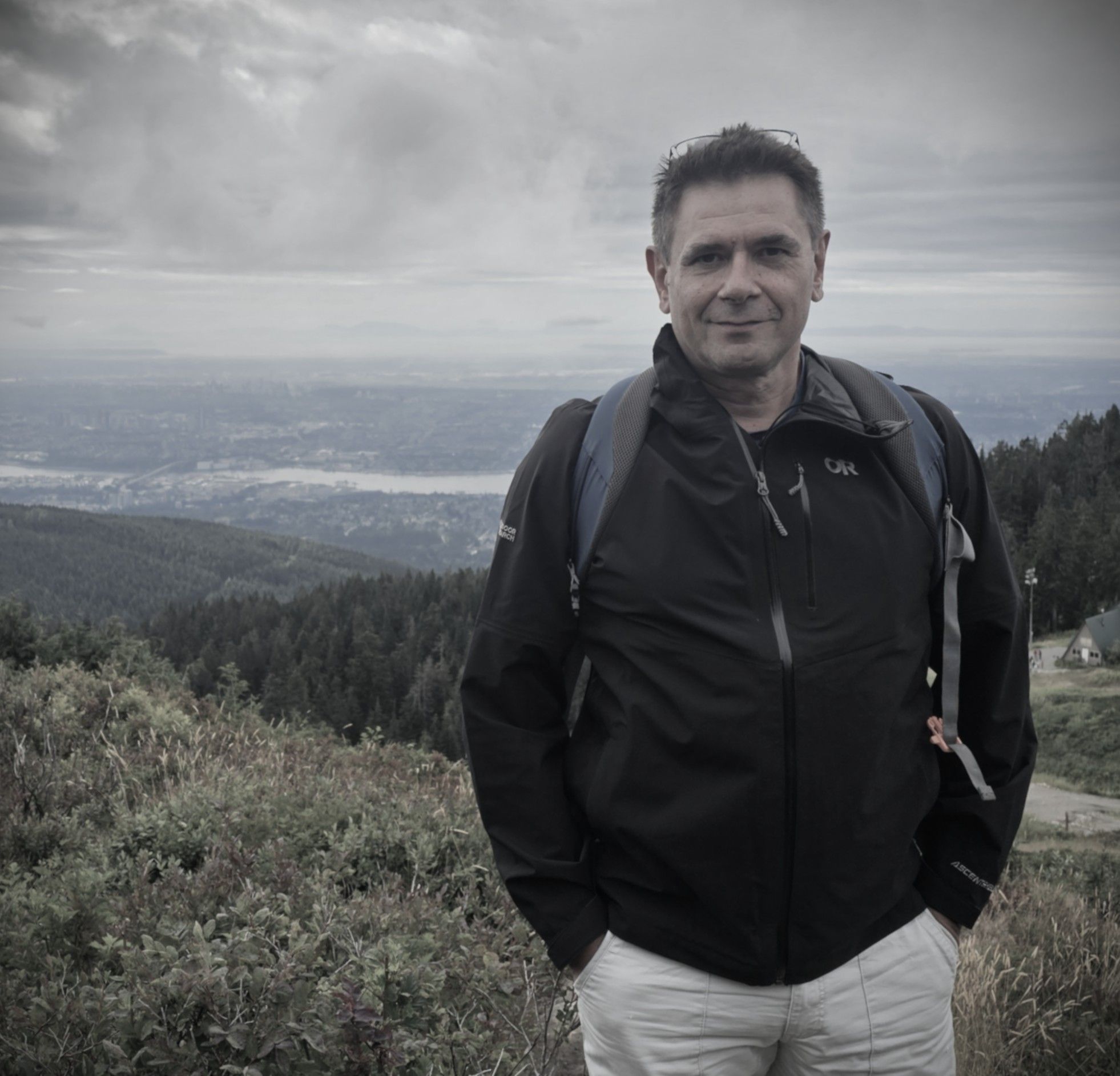The author on a mountain top in walking clothes, a far-off harbour and its water behind him.